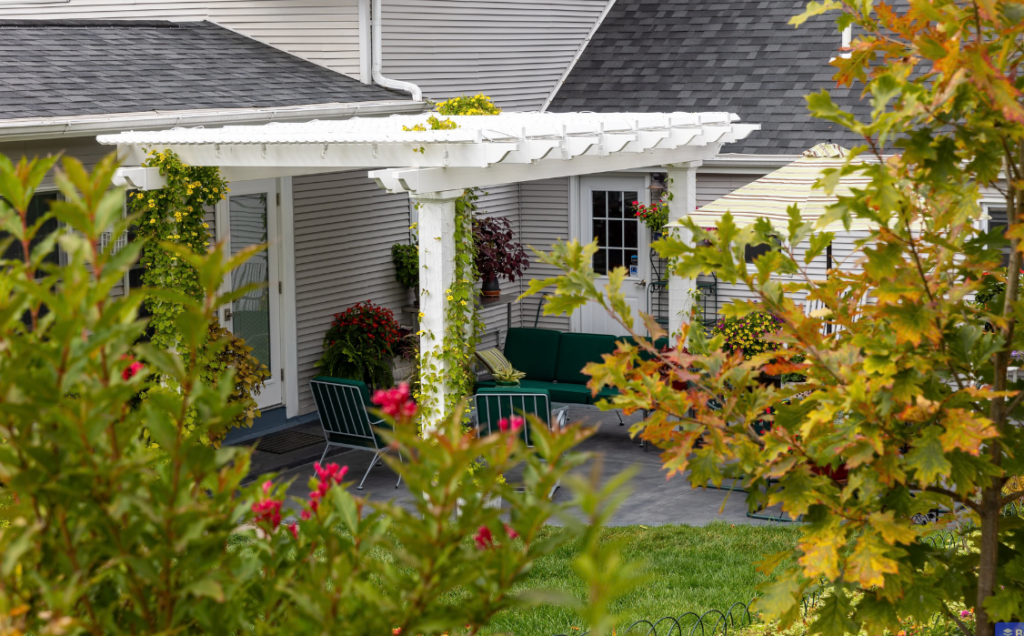 White outdoor pergola with climbing vines, surrounded by lush landscaping and flowering plants