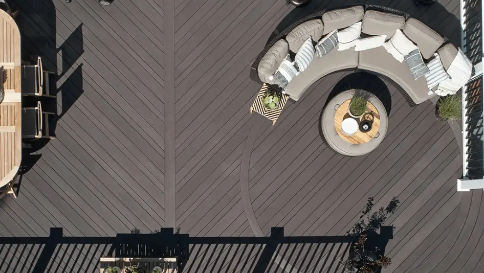 Aerial view of a patio with dark herringbone decking from a recent deck install, a curved sectional sofa with cushions and throws, a round coffee table, and shadows from a railing.