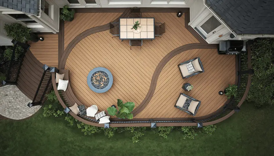 Aerial view of a modern wooden deck with curved design, featuring DIY deck materials, a fire pit with surrounding chairs, a dining table, lounge seating, potted plants, and black railing, bordered by green grass and shrubs.