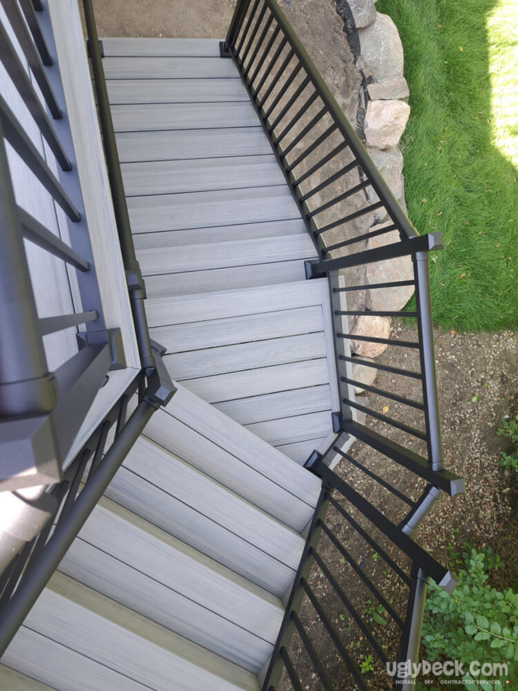 View from above of an outdoor staircase with gray composite decking and black metal railings, expertly crafted by a deck builder, leading down to a garden area with grass, rocks, and soil.