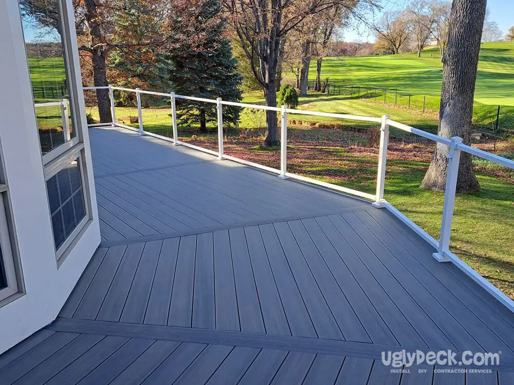 A spacious gray composite deck, built with quality DIY deck materials, features white railings and a chevron pattern. Overlooking a green lawn and trees on a sunny day, this inviting space is perfect for relaxing outdoors.