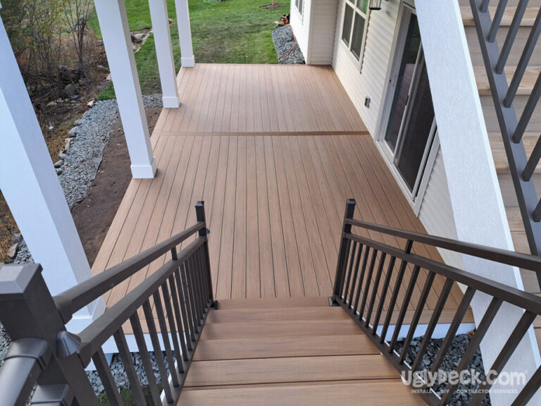 View from the top of a staircase leading down to a spacious, light brown composite deck—perfect for any DIY deck materials enthusiast—attached to a house with white columns and metal railings, overlooking a green yard.