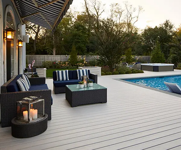Modern backyard patio with wicker furniture, striped cushions, a glass coffee table, and candles in lanterns sits beside a swimming pool. Trees and greenery surround the area at sunset—perfect inspiration if you're planning a DIY deck install.