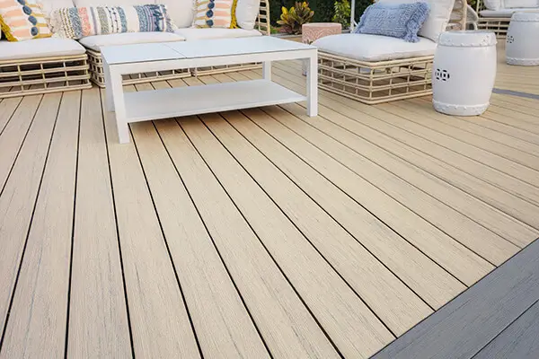Close-up of a light wood-look composite deck, perfect for DIY deck materials, with a white coffee table, wicker outdoor sofas with colorful cushions, and a white ceramic garden stool in a relaxing patio setting.