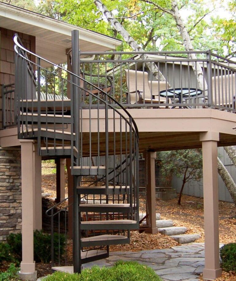 A metal spiral staircase leads up to a raised wooden deck, crafted with quality DIY deck materials and featuring a railing and outdoor chairs, surrounded by trees and a landscaped yard.
