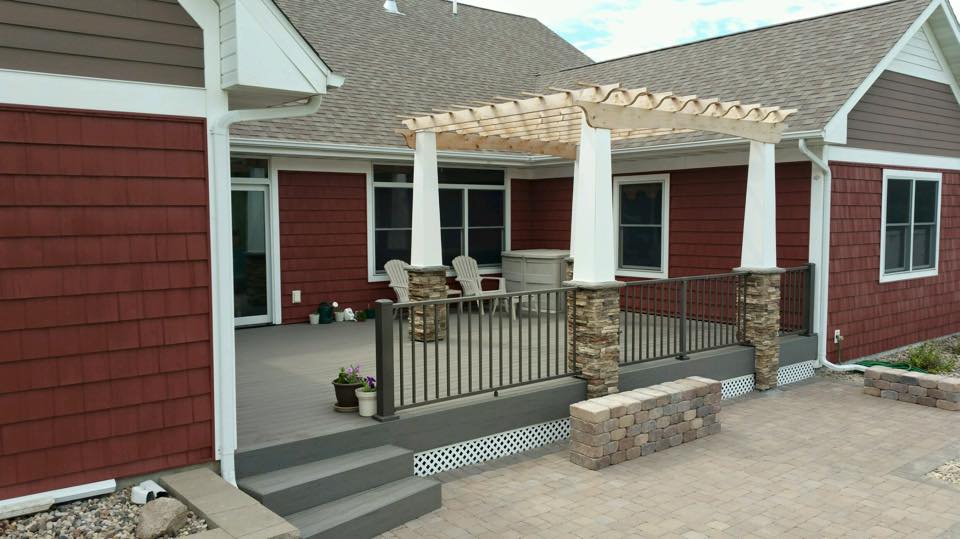 A backyard patio with red siding, a wooden pergola, two white chairs, a white table, stone pillars, black railing, potted plants, and a paved area bordered by a low stone wall—perfect inspiration for your next DIY deck materials project.
