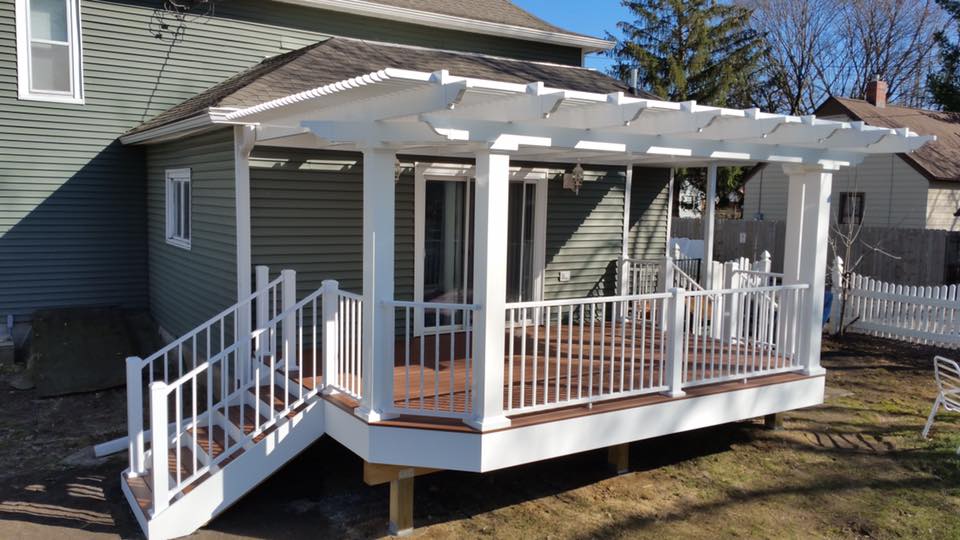 A white pergola with railings and wooden floor extends from a green house. Perfect for any DIY deck materials project, the elevated deck features stairs on both sides and sunlight casting shadows onto the sturdy posts.