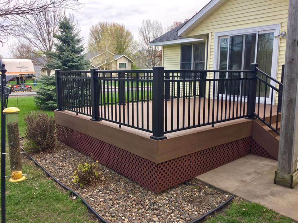 A raised wooden deck installed with black railings is attached to a yellow house. Featuring brown lattice skirting, this deck overlooks a grassy yard with trees and shrubs. A sliding glass door leads inside—perfect inspiration for your next DIY deck materials project.