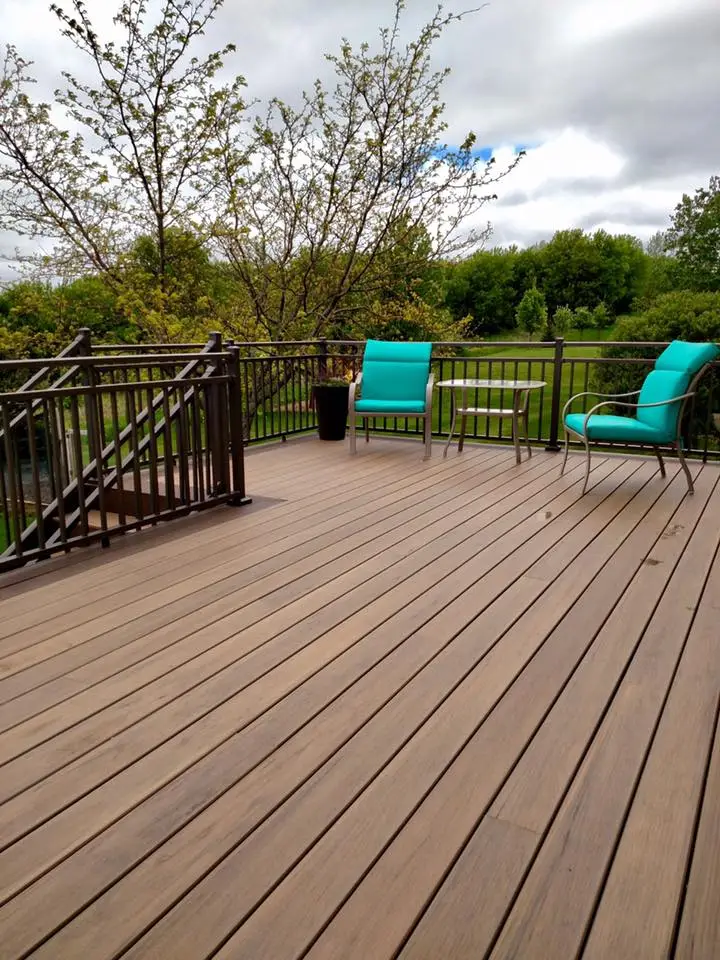 A spacious wooden deck, expertly crafted by a skilled deck builder, features dark railings, two turquoise cushioned chairs, and a small black pot overlooking a lush green lawn and trees under a cloudy sky.