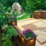 A wooden deck with geometric patterns, built using quality DIY deck materials, is surrounded by railings and flower boxes filled with colorful blooms. It overlooks a lush garden with a stone outdoor fireplace and green trees in the background.