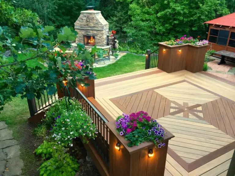 A wooden deck with geometric patterns, built using quality DIY deck materials, is surrounded by railings and flower boxes filled with colorful blooms. It overlooks a lush garden with a stone outdoor fireplace and green trees in the background.
