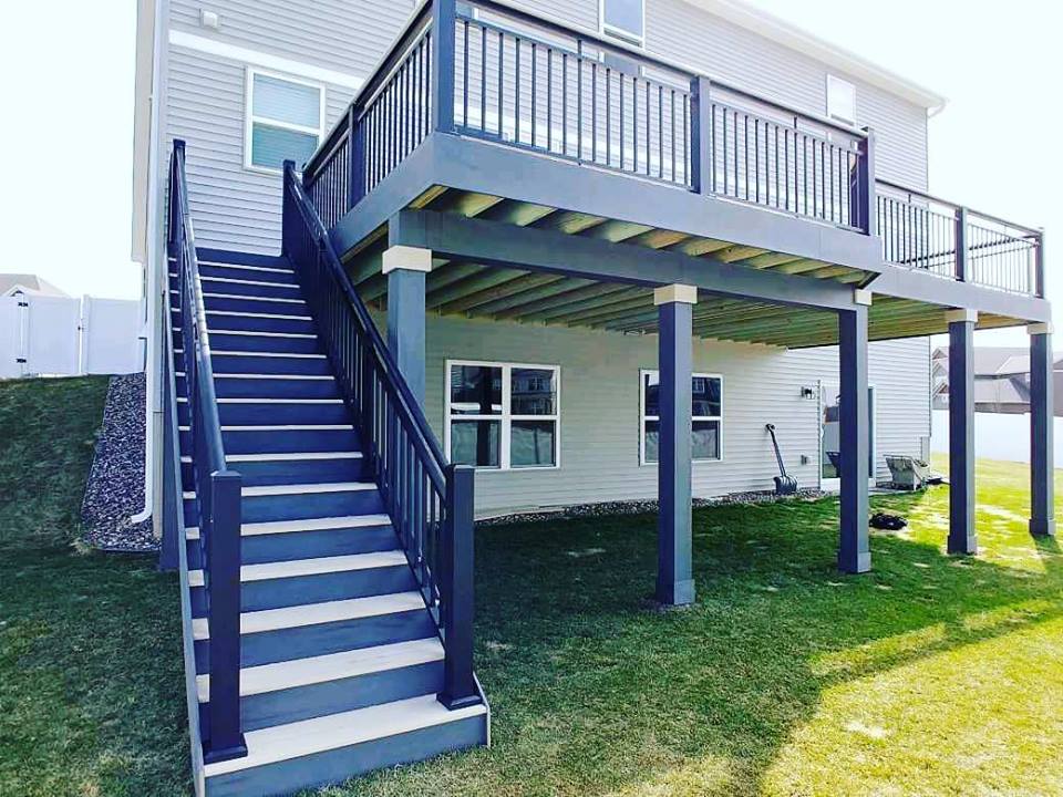A two-story house with a large raised deck built using quality DIY Deck Materials, featuring black railings and gray steps leading down to a grassy backyard. The sturdy deck is supported by pillars, with windows on the lower level.