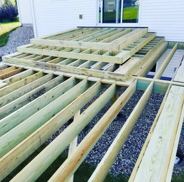 Partially constructed wooden deck frame attached to a white house, with exposed beams and joists over a gravel and grass yard. The decking boards have not yet been installed—ideal for those seeking DIY deck materials for their next deck install.