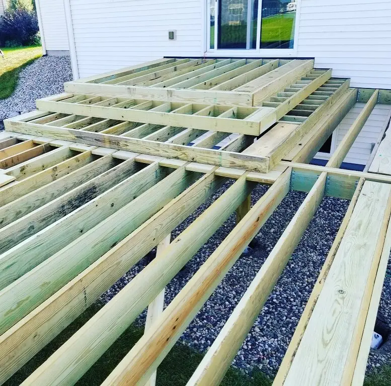 Partially constructed wooden deck frame attached to a white house, with exposed beams and joists over a gravel and grass yard. The decking boards have not yet been installed—ideal for those seeking DIY deck materials for their next deck install.