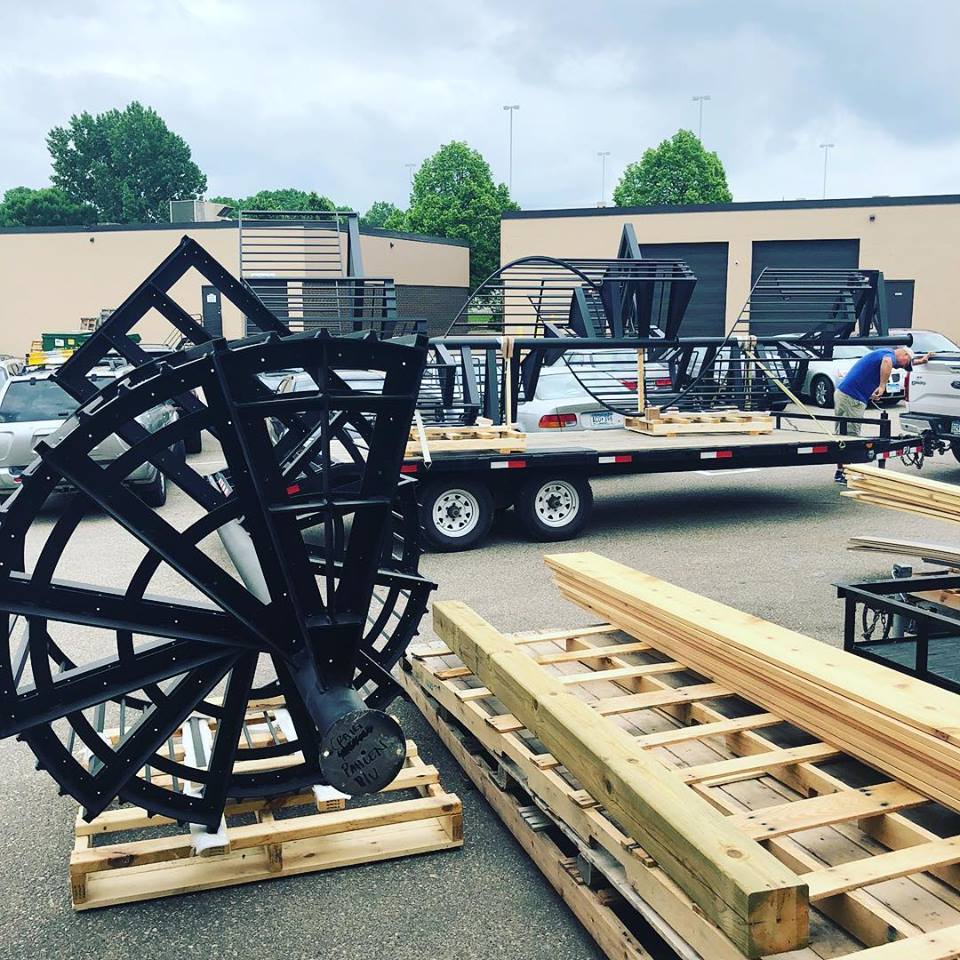 Metal and wooden construction materials, perfect for a deck builder or those seeking DIY deck materials, are loaded on pallets and a flatbed trailer in a parking lot outside an industrial building.