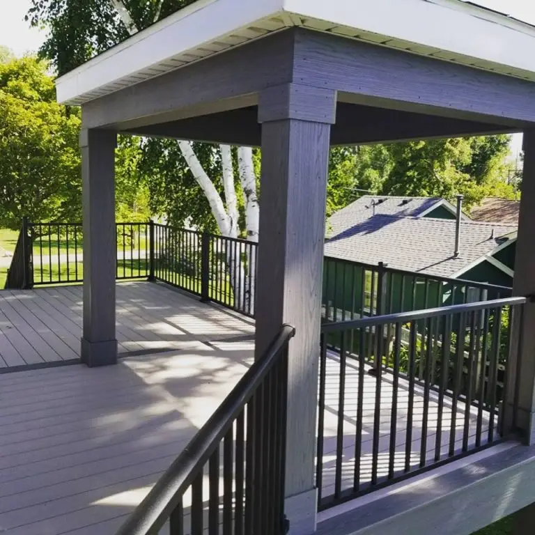 A modern outdoor deck install with gray flooring and railings, featuring a covered pergola structure with square columns. Trees and nearby houses are visible in the background under a sunny sky.