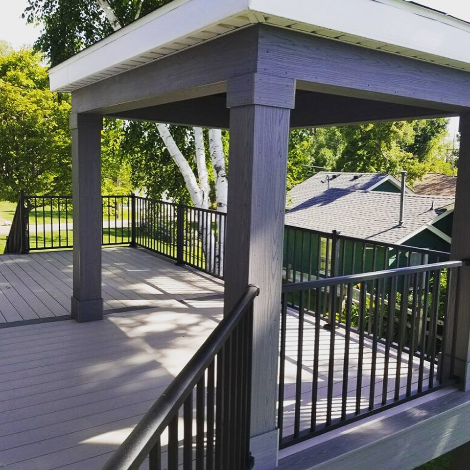 A modern outdoor deck install with gray flooring and railings, featuring a covered pergola structure with square columns. Trees and nearby houses are visible in the background under a sunny sky.