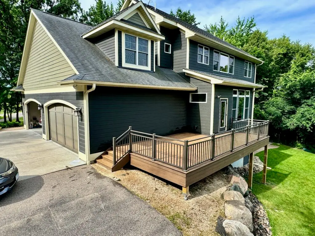 A two-story dark gray house with white trim, attached garage, and a raised wooden deck installed by a professional deck builder. The deck overlooks a grassy yard bordered by trees and landscaping rocks.