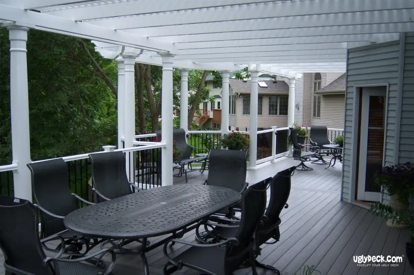 A spacious deck by a trusted Deck Builder features a white pergola, black metal dining set, and railing with views of a leafy suburban neighborhood, all attached to a light gray house.