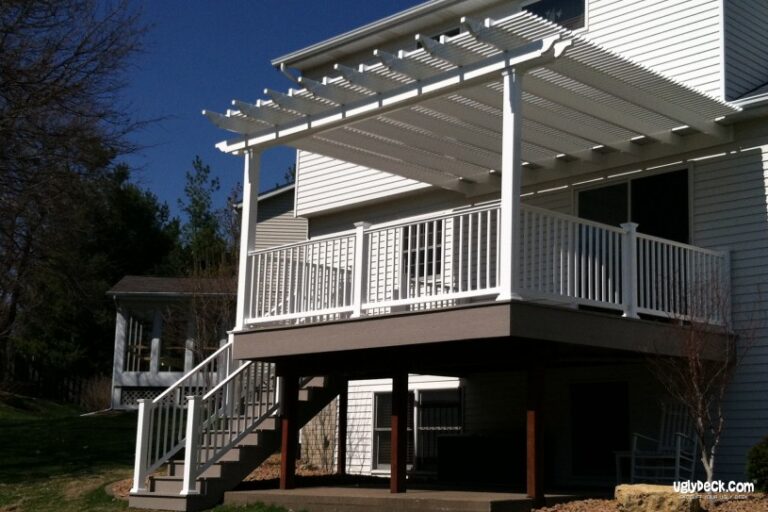 A two-story white house features a raised wooden deck built with quality DIY Deck Materials, white railings, and a white pergola above. Stairs lead from the yard to the deck, with extra space available underneath.