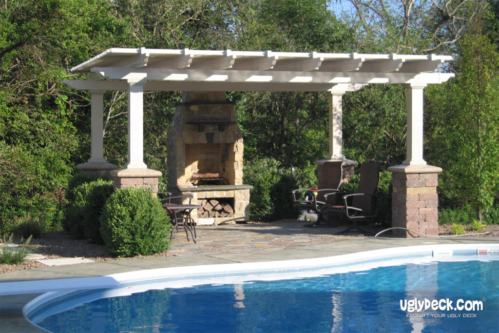 A pergola with stone columns and a built-in outdoor fireplace sits on a stone patio surrounded by greenery, next to a curved swimming pool. Patio chairs are arranged under the pergola. This inviting space highlights expert deck install by uglydeck.com.