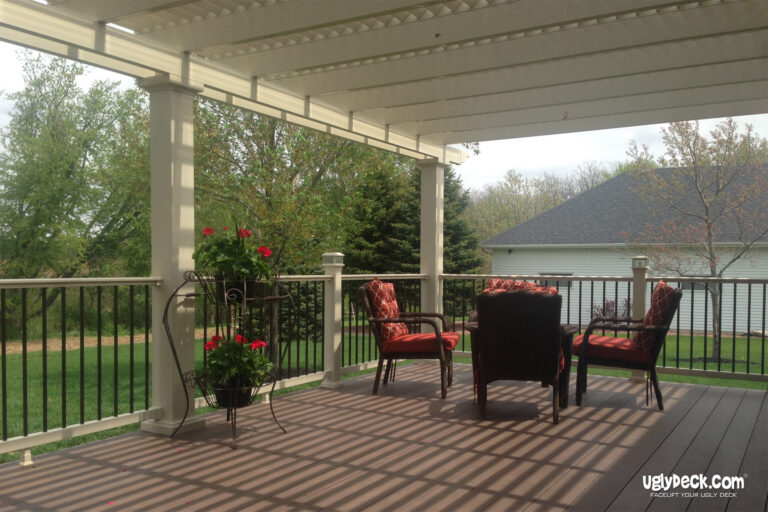 A covered deck install features dark wood flooring, black railings, and four cushioned chairs around a small table. Red flowers in pots decorate the space, with a grassy yard and trees visible in the background.