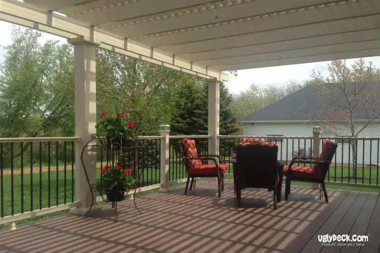 A covered deck install features dark wood flooring, black railings, and four cushioned chairs around a small table. Red flowers in pots decorate the space, with a grassy yard and trees visible in the background.
