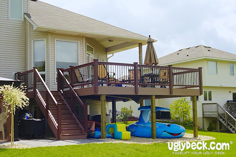 A raised backyard deck with brown railings and stairs, built by a professional Deck Builder, features outdoor furniture and an umbrella. Beneath the deck are a blue inflatable pool toy and children's play items in a suburban neighborhood.
