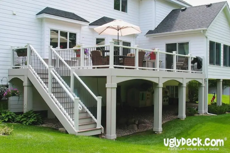 A two-level deck attached to a white house features DIY Deck Materials, patio furniture, an umbrella, and white railings. Stairs lead to the landscaped lawn and a small playhouse under the deck. The UglyDeck.com logo is visible.
