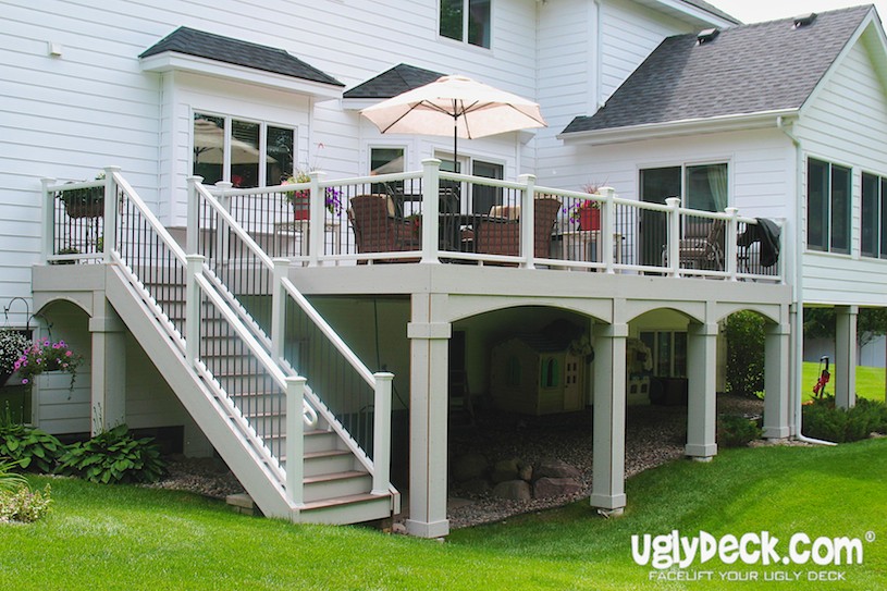 A raised deck with white railings and stairs attached to a white house, featuring outdoor furniture and an umbrella. Underneath is a playhouse and mulched area with rocks—perfect inspiration for your next Deck Builder or DIY Deck Materials project. UglyDeck.com logo bottom right.