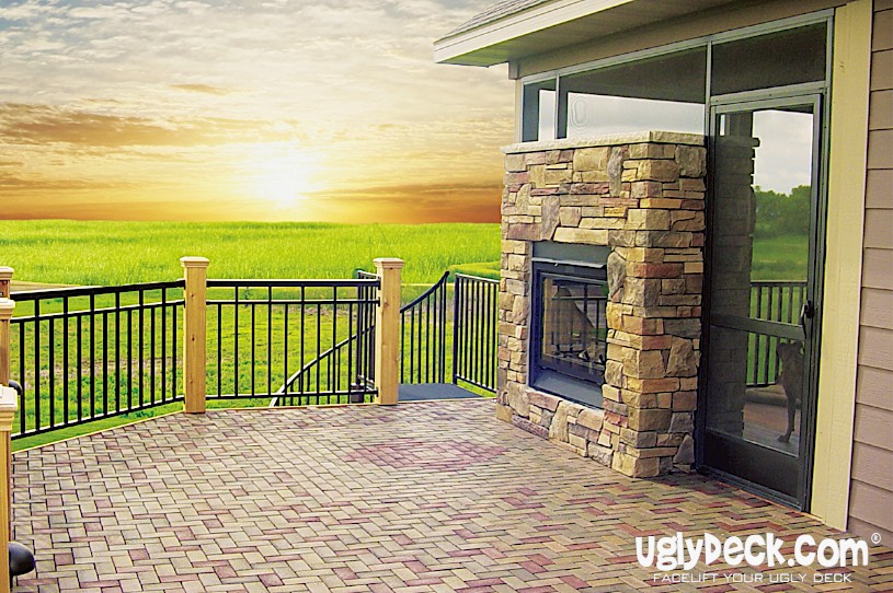 A patio with a stone fireplace, brick flooring, and black railing overlooks a grassy field at sunset. The sky is partly cloudy, adding to the peaceful scene. “uglydeck.com,” a trusted deck builder, brands the image in the bottom right corner.