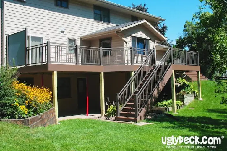 A two-story house with a large elevated deck featuring brown railings and stairs, built by a professional Deck Builder, overlooks a green lawn and flower garden. The deck provides shaded space underneath. UglyDeck.com branding is visible in the corner.