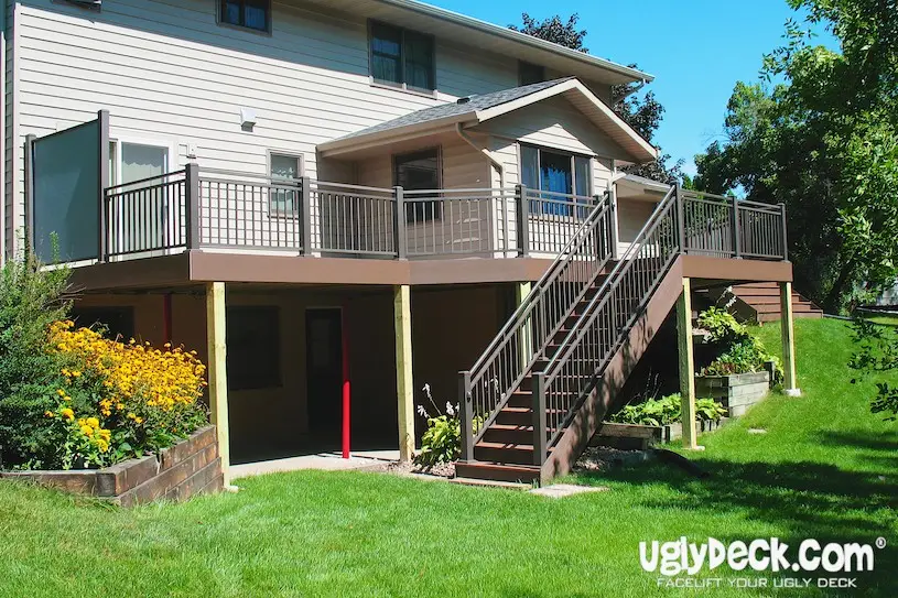 A two-story house with a large elevated deck featuring brown railings and stairs, built by a professional Deck Builder, overlooks a green lawn and flower garden. The deck provides shaded space underneath. UglyDeck.com branding is visible in the corner.