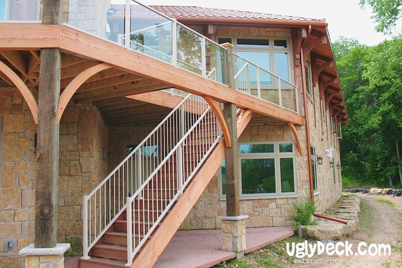 A two-story stone house features a wooden deck with glass railings, showcasing quality DIY Deck Materials. A staircase leads to the lower level, all surrounded by greenery and large windows. The logo uglydeck.com appears in the bottom right corner.