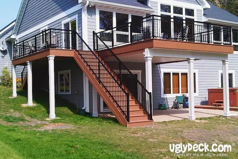 Two-story gray house with a large elevated deck featuring black railings, stairs leading to a patio, and white column supports. Ideal for those seeking DIY deck materials or considering a professional deck builder for their next deck install.