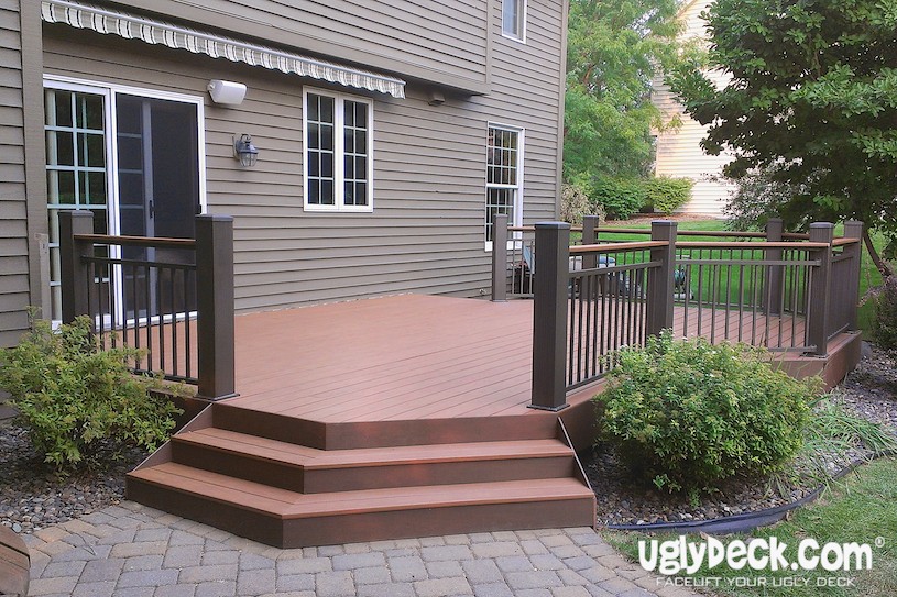 A modern wooden deck with black railings and three steps attached to a tan house, surrounded by landscaping and a brick patio. Expert deck builder craftsmanship is shown, with the uglydeck.com logo branded in the corner.