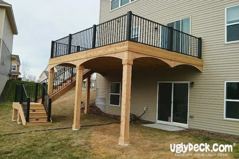 A two-story house with a wooden elevated deck, crafted by an expert Deck Builder, features black railings and a staircase leading to the yard. The space beneath the deck is open, and uglyDeck.com appears in the bottom right corner.
