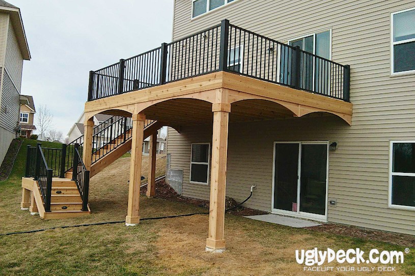 A two-story house with a wooden elevated deck, crafted by an expert Deck Builder, features black railings and a staircase leading to the yard. The space beneath the deck is open, and uglyDeck.com appears in the bottom right corner.