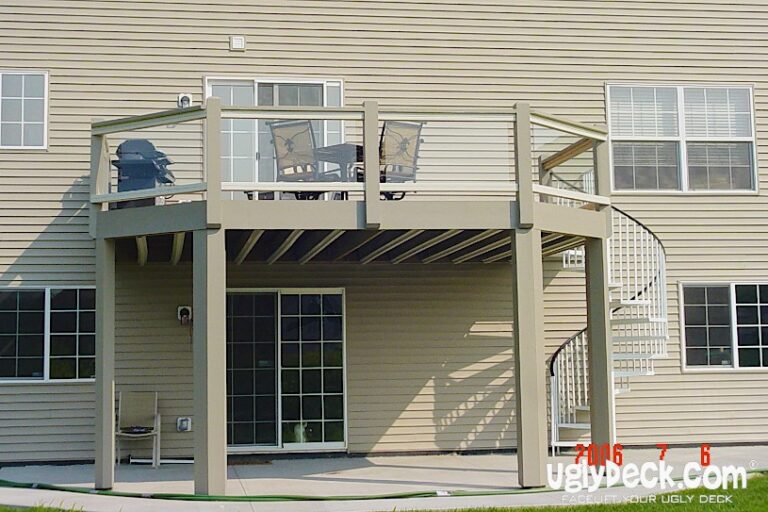 A beige two-story house with a raised deck, expertly crafted by a Deck Builder, features glass railings and a spiral staircase leading to a patio area. Patio furniture sits atop the deck, with the structure and underside clearly visible.