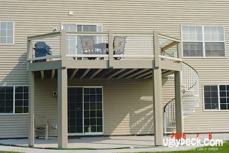 A beige two-story house with a raised deck, expertly crafted by a Deck Builder, features glass railings and a spiral staircase leading to a patio area. Patio furniture sits atop the deck, with the structure and underside clearly visible.