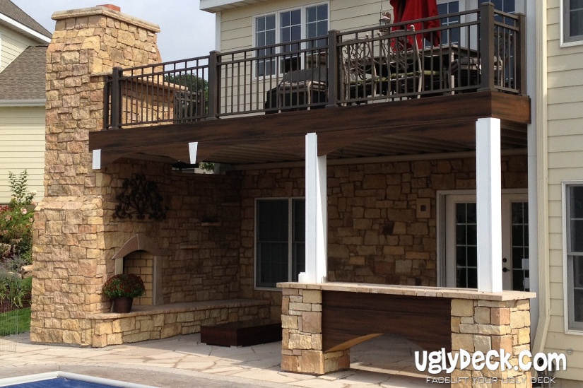 A two-story patio with a wooden deck install and railing above, stone fireplace and seating area below, plus a stone bar beside the swimming pool. The house exterior is light-colored with large windows. UglyDeck.com is visible in the corner.