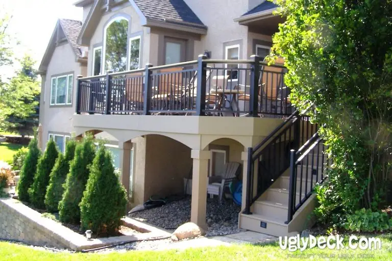 A two-story house with a raised deck featuring black railings and stairs by a skilled Deck Builder. Beneath the deck is a shaded sitting area with chairs. Neatly trimmed bushes and a stone-bordered garden line the yard. uglydeck.com is visible in the corner.