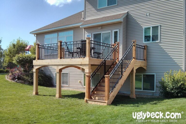A two-story house with a raised wooden deck, built using quality DIY Deck Materials, features black metal railings and a staircase to a grassy backyard. Patio furniture is on the deck, and uglydeck.com is branded in the bottom right corner.
