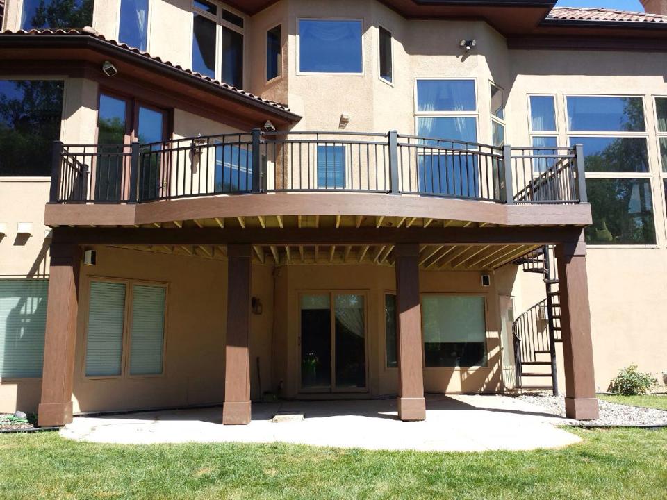 Two-story beige house with large windows and a balcony featuring black metal railings. The balcony is supported by wooden posts, and a spiral staircase built by a skilled deck builder descends to a patio and green lawn.