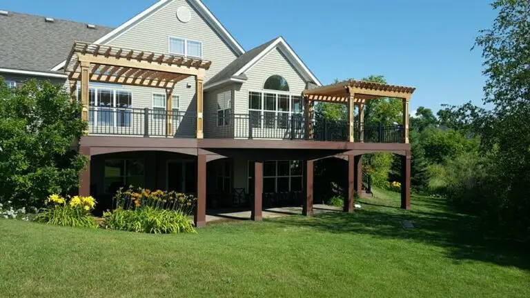 A two-story house with beige siding and large windows features an elevated deck install with pergolas, black railings, and a shaded patio beneath. Yellow flowers and green lawn surround the home on a sunny day.