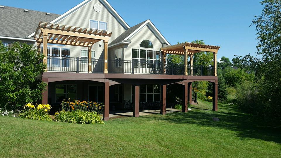 A two-story house with beige siding and large windows features an elevated deck install with pergolas, black railings, and a shaded patio beneath. Yellow flowers and green lawn surround the home on a sunny day.