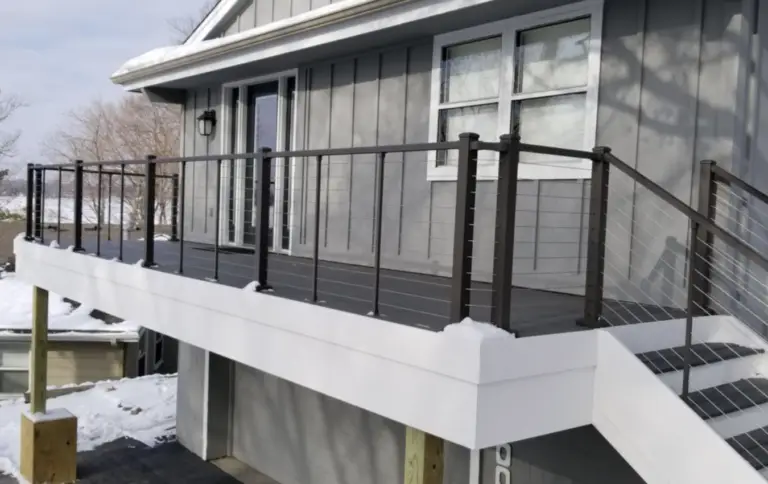 A modern gray house with a raised deck featuring dark railings, cable wires, and white trim. Snow covers the ground and deck railing. Perfect for a DIY Deck Install, stairs lead down from the deck to the driveway below.