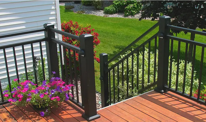 A wooden deck built with quality DIY deck materials and black metal railings overlooks a green lawn and garden. A planter with bright pink and purple flowers sits on the deck, with bushes and shrubs growing alongside the railing.