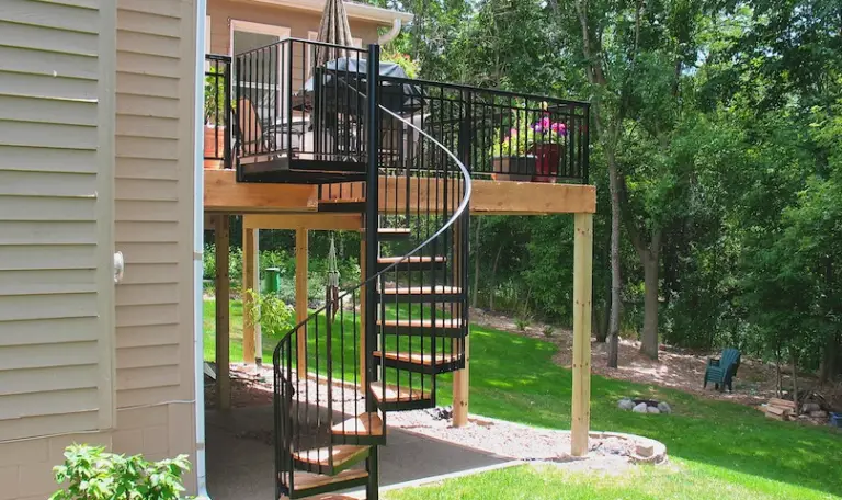 A wooden deck built by a professional Deck Builder features black metal railings and a spiral staircase leading to a landscaped backyard. Patio furniture and potted plants add charm to this inviting outdoor space.