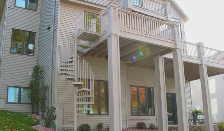 A beige house features a raised wooden deck built with DIY deck materials, white railings, and a spiral metal staircase leading to a landscaped backyard with large windows on the lower level.